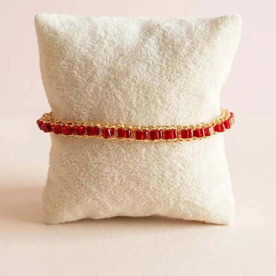 A gold chain bracelet with rectangular red gemstones, displayed on a textured cream pillow against a soft pink background.