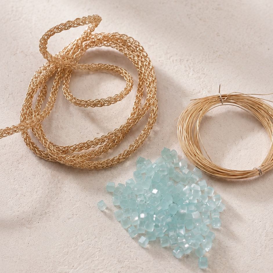 Gold wire coiled into two loops and a pile of light blue faceted glass beads on a textured beige surface with soft shadows.