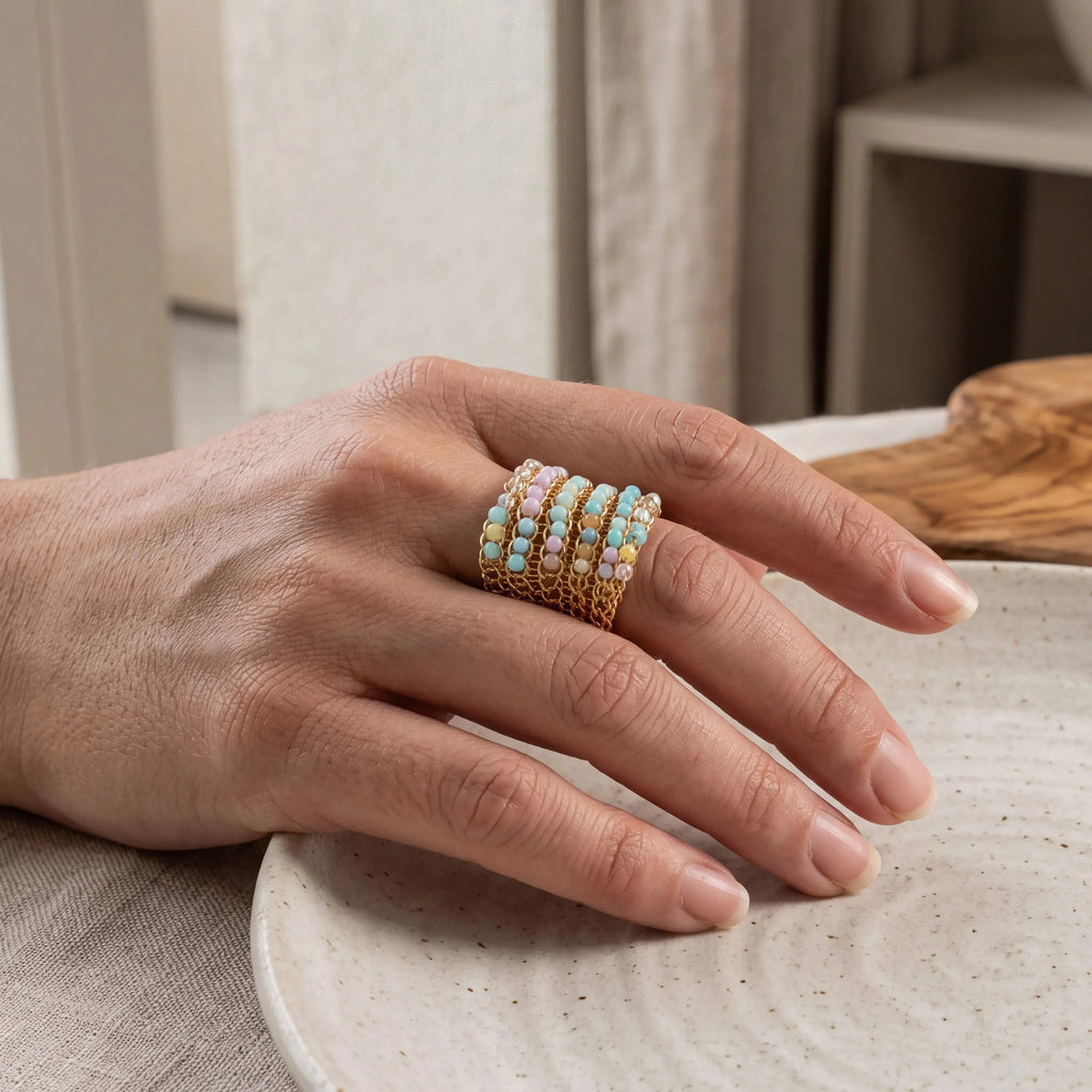 A close-up of a hand wearing a stack of gold rings adorned with small pastel-colored stones, resting on a textured ceramic plate in a softly lit indoor setting.