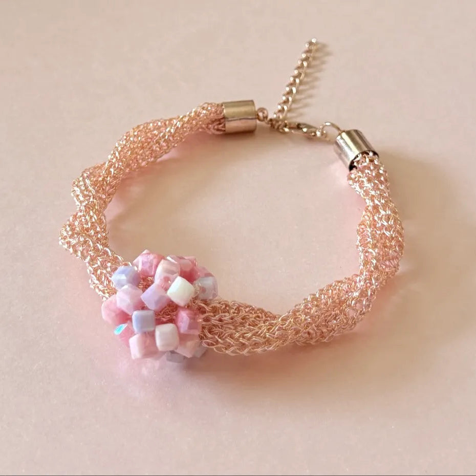A delicate mesh bracelet in a soft peach-pink tone with a central cluster of pastel pink, lavender, and white beads, finished with gold-toned metal caps and a chain extender on a light beige background.