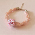 A delicate mesh bracelet in a soft peach-pink tone with a central cluster of pastel pink, lavender, and white beads, finished with gold-toned metal caps and a chain extender on a light beige background.