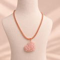 A rose gold chain necklace with a heart-shaped pendant made of light pink seed beads, displayed on a cream fabric mannequin bust against a soft beige background.