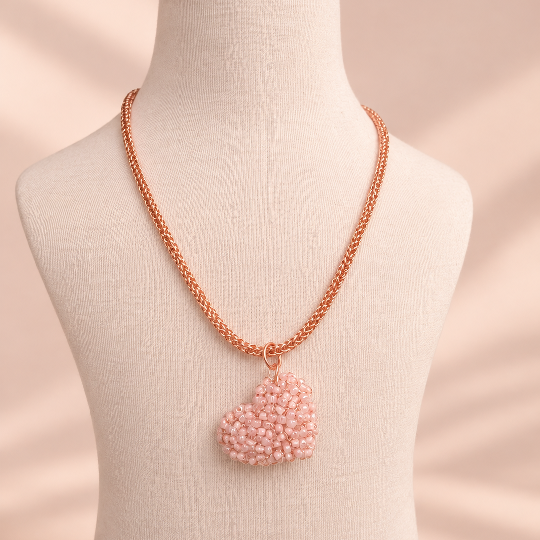 A rose gold chain necklace with a heart-shaped pendant made of light pink seed beads, displayed on a cream fabric mannequin bust against a soft beige background.