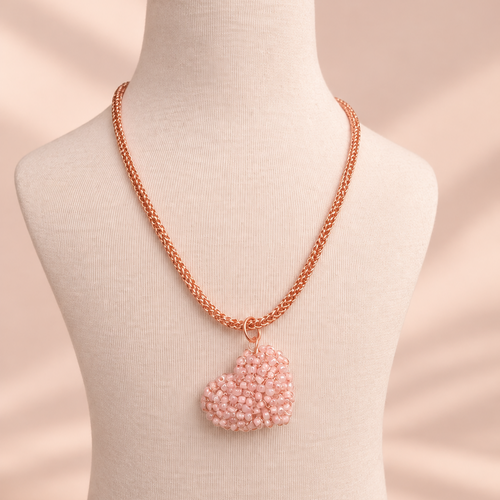 A rose gold chain necklace with a heart-shaped pendant made of light pink seed beads, displayed on a cream fabric mannequin bust against a soft beige background.