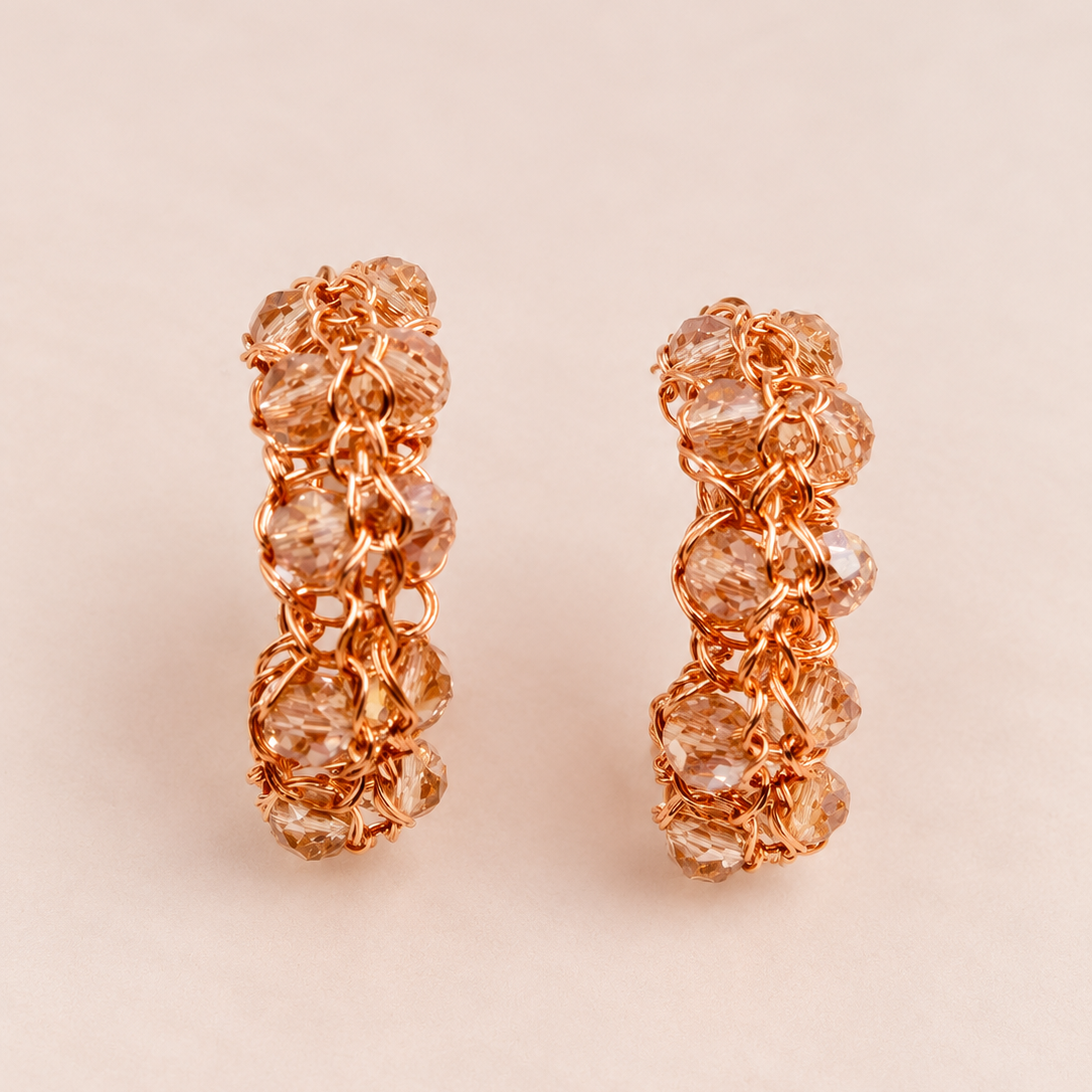 A pair of rose gold hoop earrings featuring a textured, beaded floral pattern with clear faceted crystals, set against a solid pale pink background.
