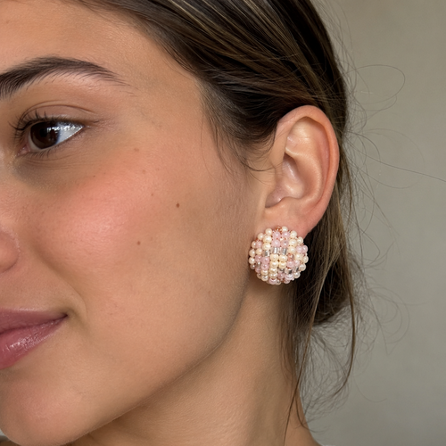 A close-up of a woman wearing a round, textured earring made of small pale pink and white beads, set against a neutral beige background.
