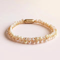 A double-strand beaded bracelet featuring white faux pearls and clear faceted crystals woven with gold-toned metal links, finished with a gold capsule clasp, set against a soft pink background.