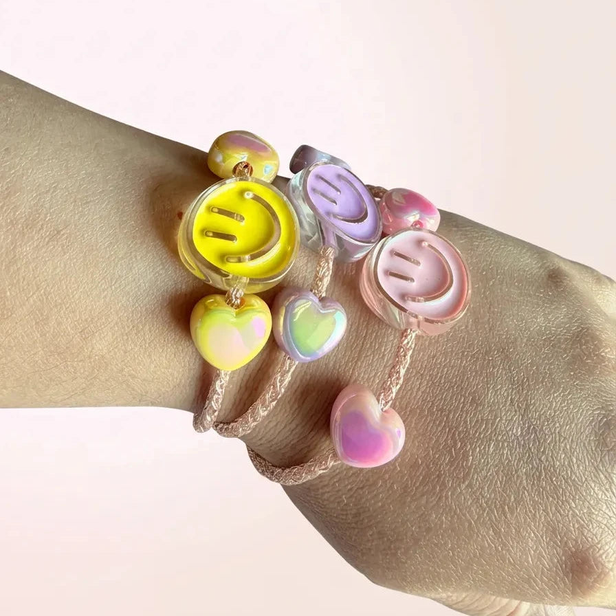 Three pastel smiley face charm bracelets on a person's wrist, featuring yellow, lavender, and pink charms with iridescent heart accents, set against a soft pink background.